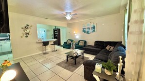 Una sala de estar con un sofá, dos sillones, una mesa de centro, un televisor y una cocina al fondo.