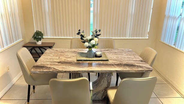 A dining area with a marble table, beige chairs, a vase with flowers, and a plant on a side table.