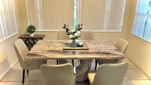 Een eetgedeelte met een marmeren tafel, beige stoelen, een vaas met bloemen en een plant op een bijzettafeltje.