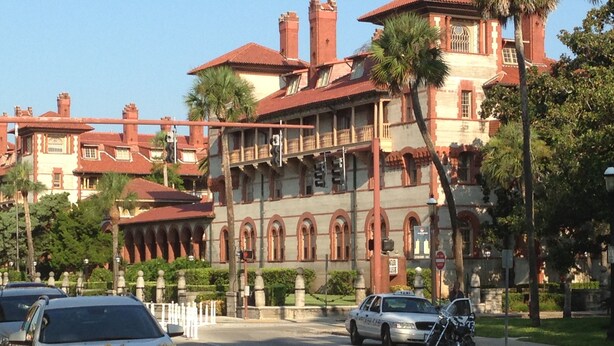 A historic building with arched windows and a balcony, surrounded by palm trees and parked cars.
