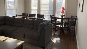 A living room with a sectional sofa, a dining table with chairs, and a kitchen area with a window. - 1st Class Rentals Munson Manor VI at Chestnut 2nd Floor (Oneonta)