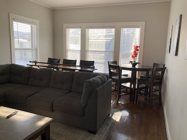 A living room with a sectional sofa, a dining table with chairs, and a kitchen area with a window.