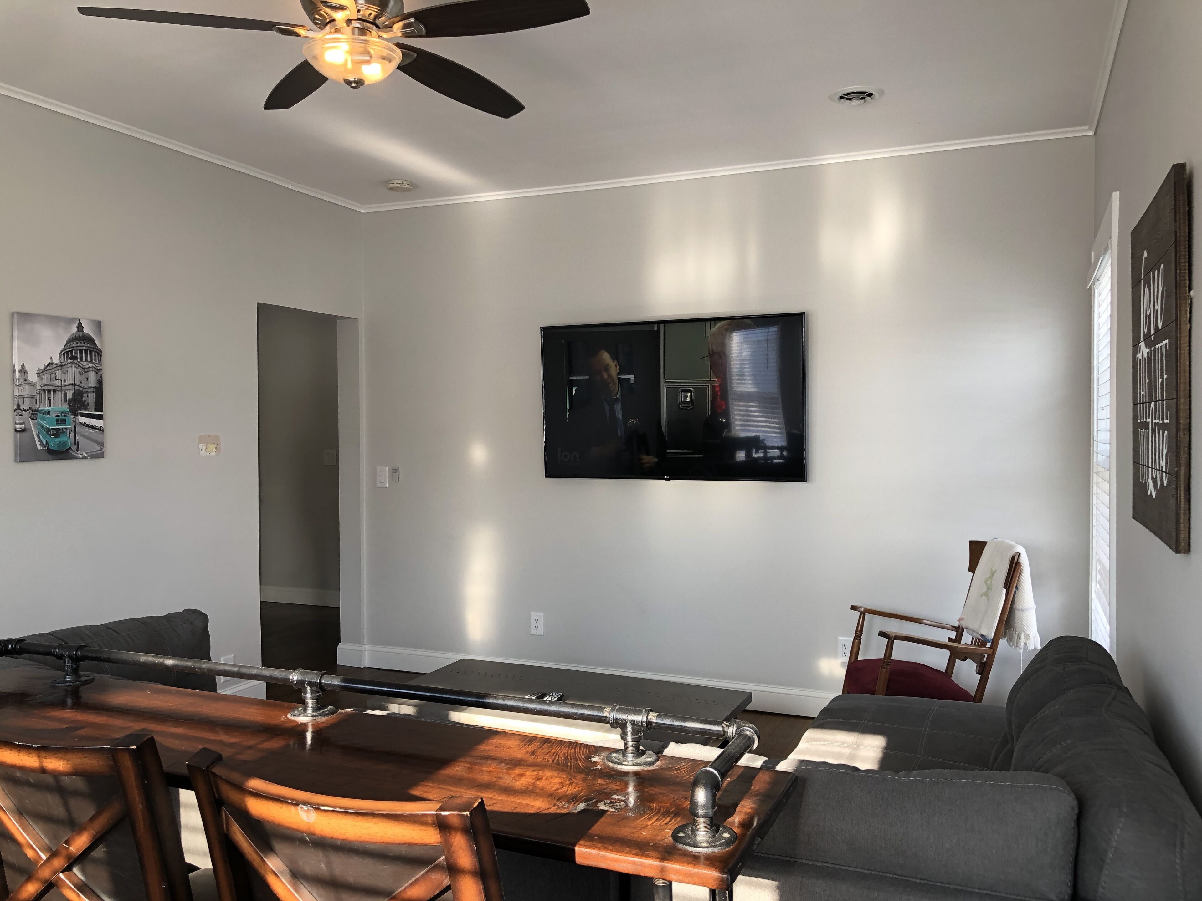 A modern living room with a dining area, a ceiling fan, a flat-screen TV, and a painting of a building.