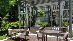 Eine Terrasse im Freien mit Holzboden, Korbmöbeln und einer Glaseinfriedung mit Blick auf ein Gebäude und Grünflächen.