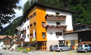 A multi-story hotel with balconies adorned with flowers, a visible sign for 'Hotel Oswald', and a parking area with cars. - Hotel Oswald (Canazei)
