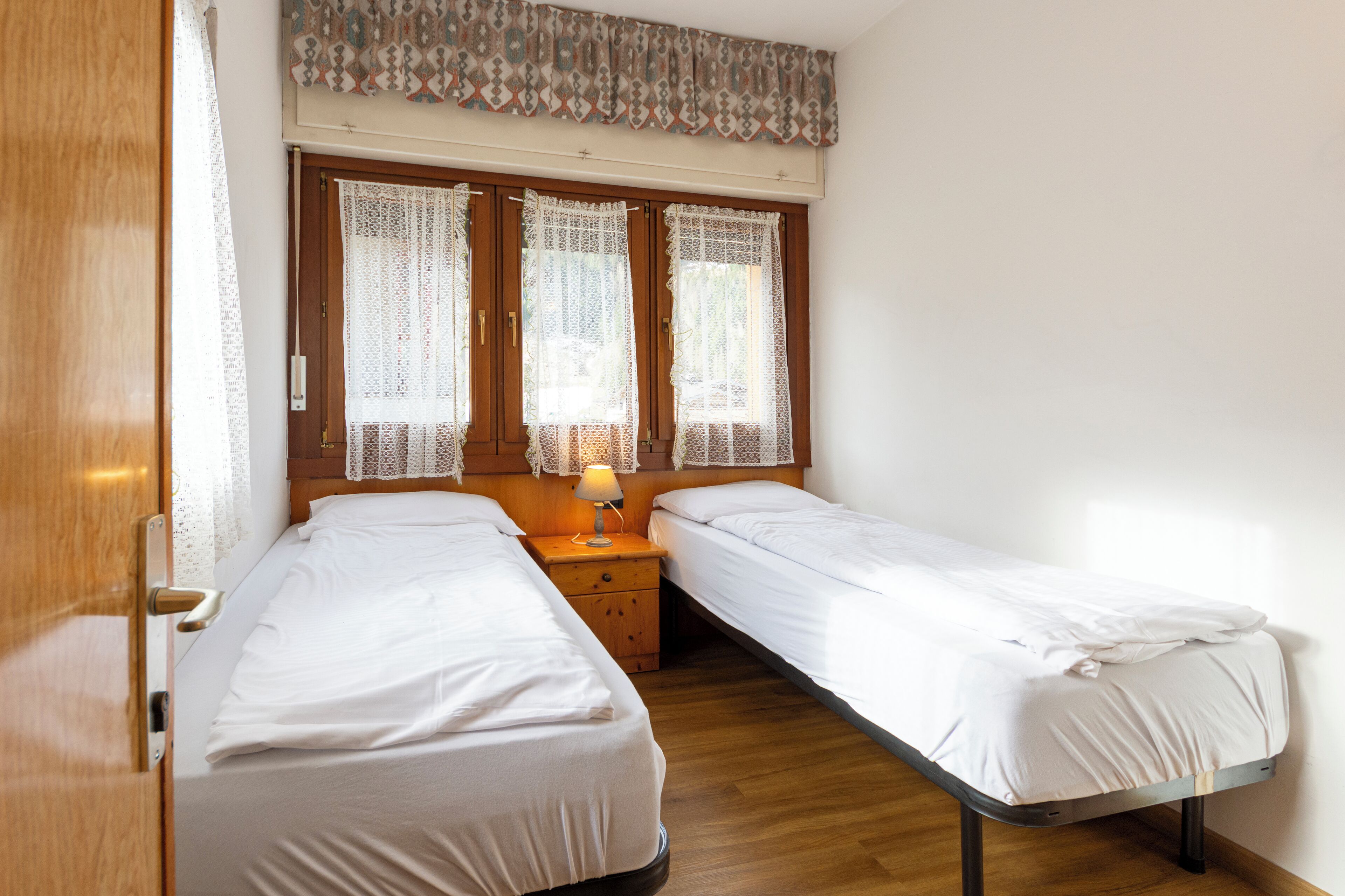 Una stanza con due letti singoli, un comodino in legno con una lampada, tende di pizzo e una porta in legno.