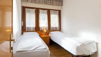 Una stanza con due letti singoli, un comodino in legno con una lampada, tende di pizzo e una porta in legno.