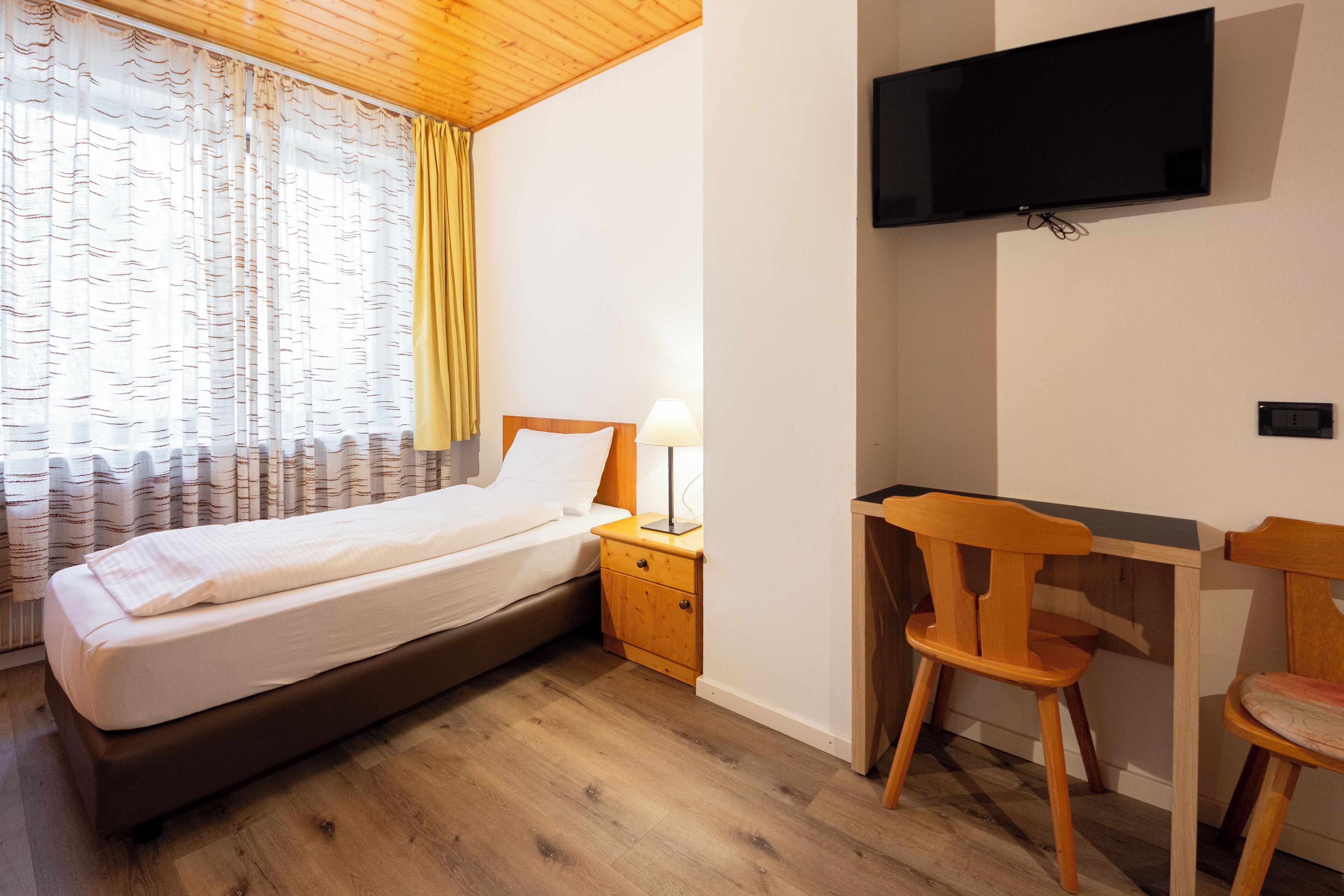A bedroom with a bed, a desk, a chair, a TV, and a window with curtains.