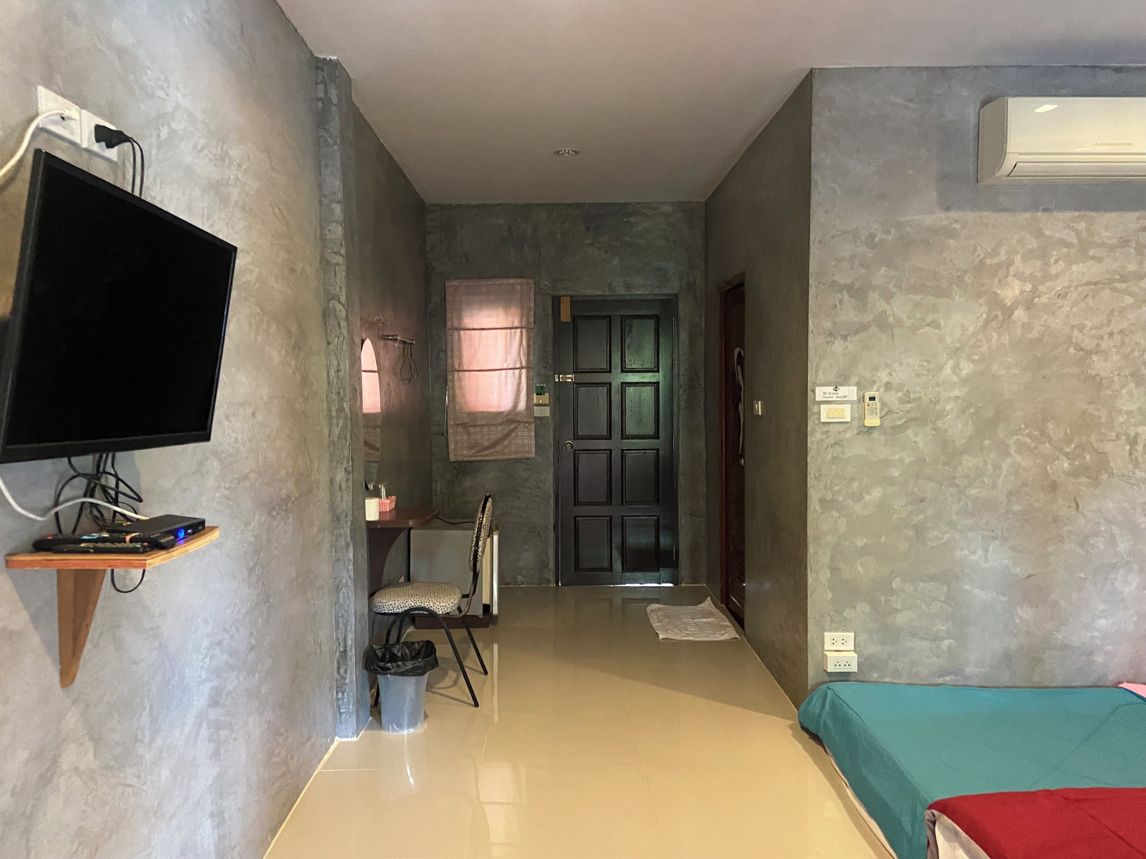 Una habitación con un televisor de pantalla plana montado en la pared, una cama con colcha verde azulada y roja, un estante de madera con dispositivos electrónicos, una silla, un cesto de basura y una puerta que da a otra habitación.