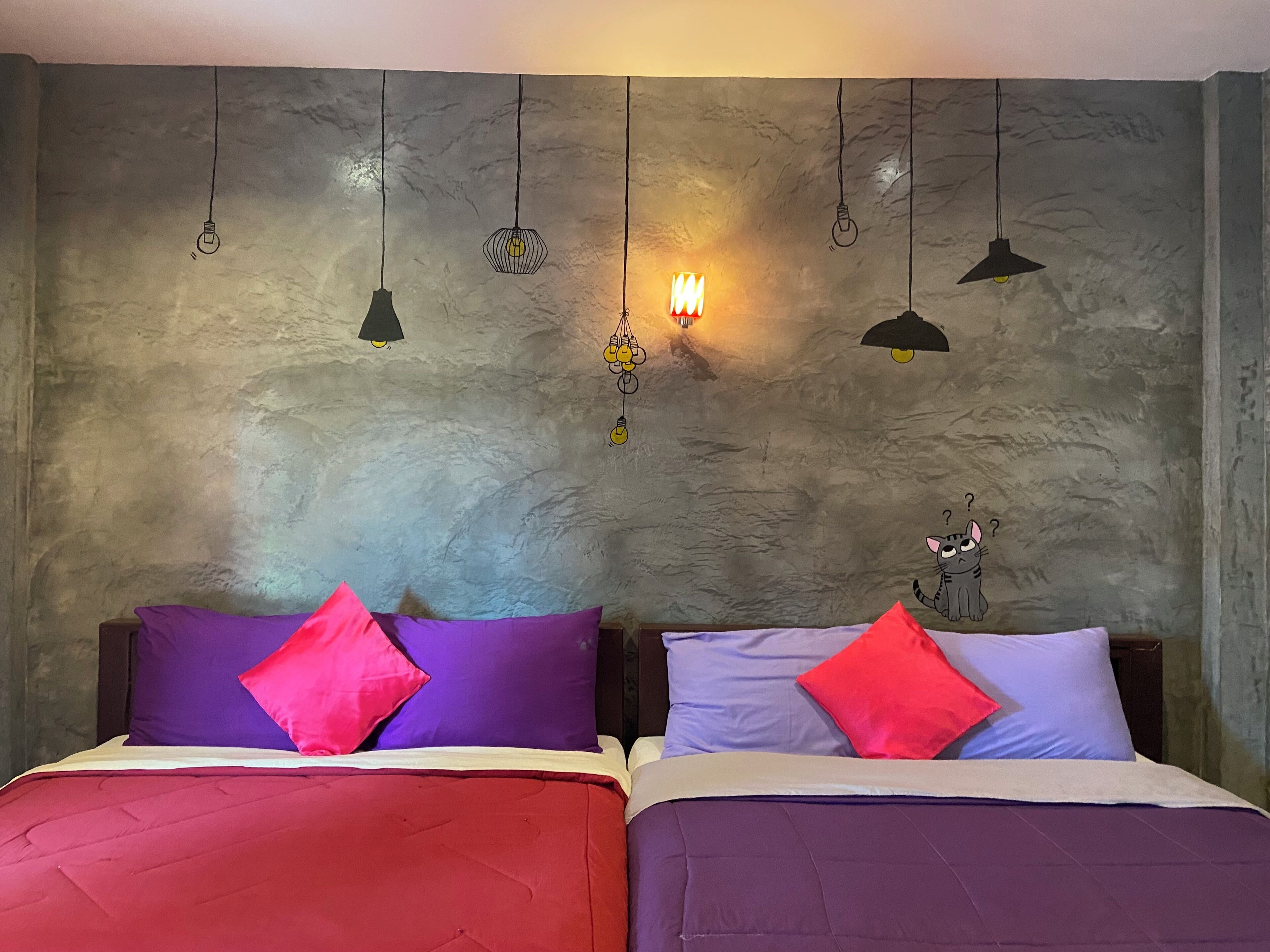 Habitación con dos camas, cada una con un edredón y almohadas de diferente color. Varias lámparas colgantes en la pared.
