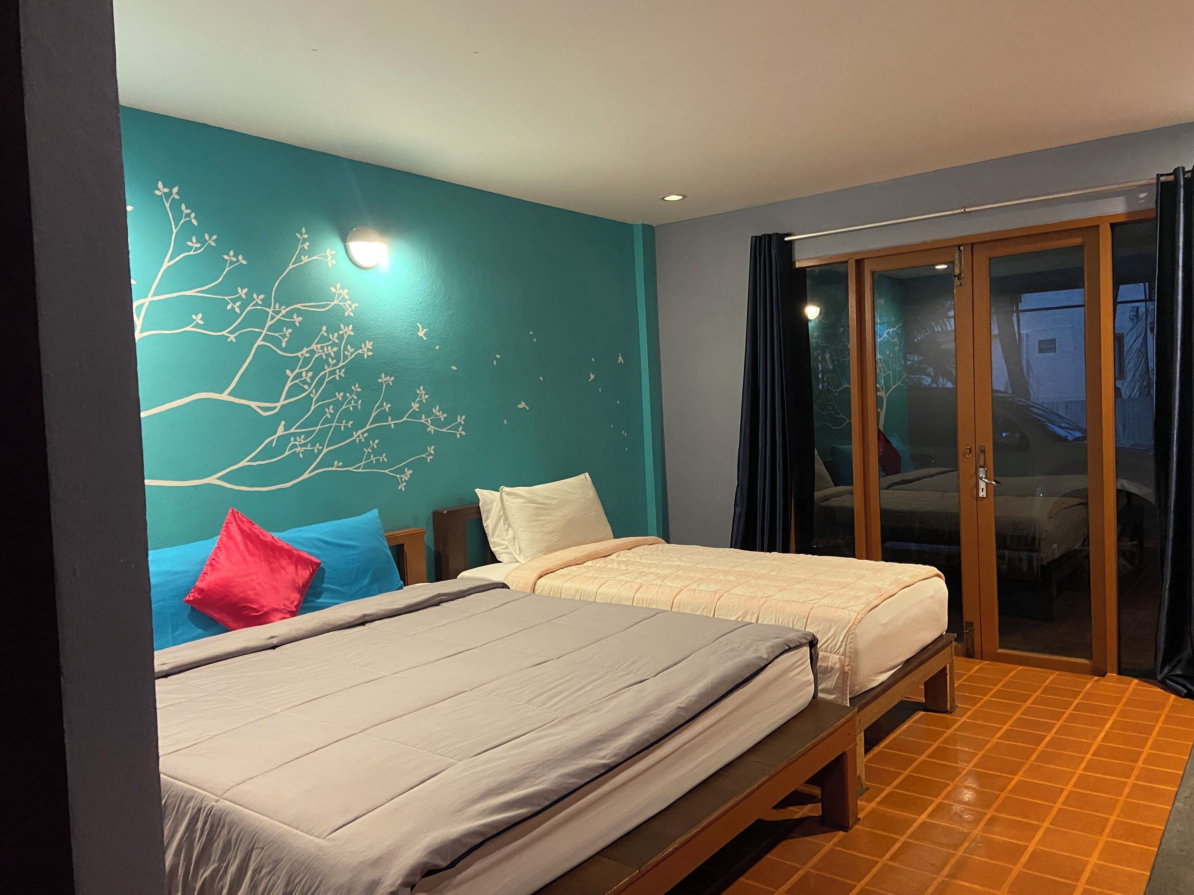 A room with two beds, a teal wall with a tree design, and a sliding glass door leading outside.