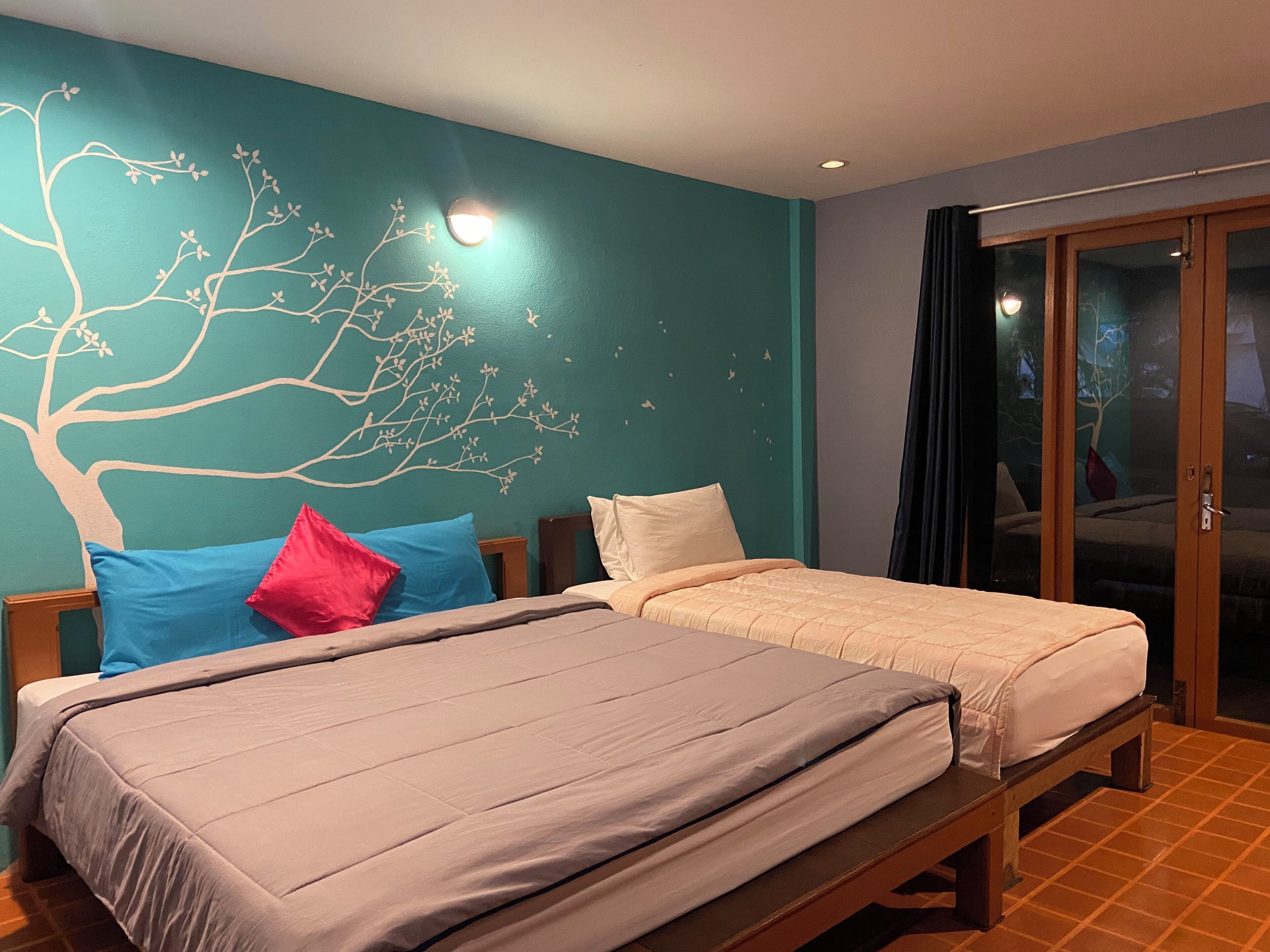 Cama doble con colcha color beige, mesita de noche con lámpara y un mural en la pared con un árbol.