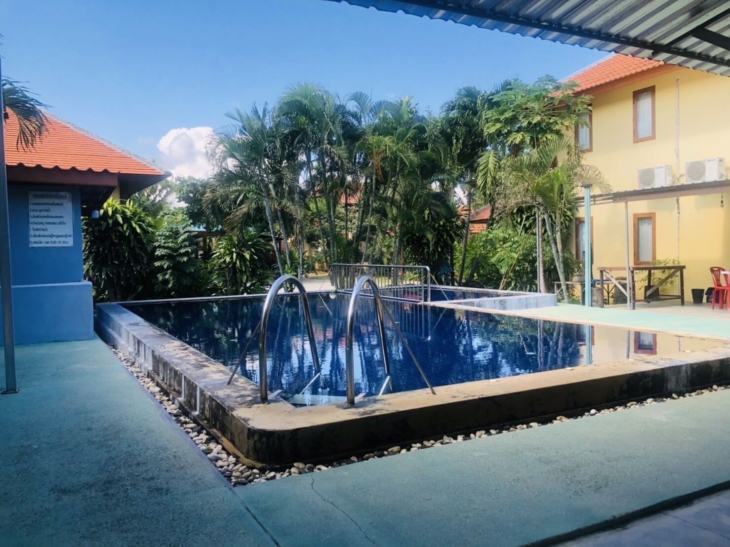 Ein Schwimmbecken mit Metallleitern, umgeben von Grünflächen und einem Gebäude mit rotem Dach.
