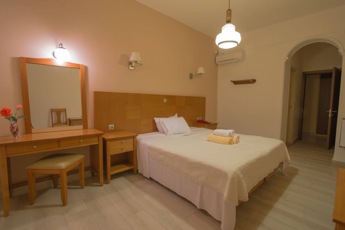Une chambre d’hôtel avec un bureau en bois, une chaise, un lit recouvert d’une courtepointe blanche, une table de chevet, un miroir et une lampe fixée au mur.