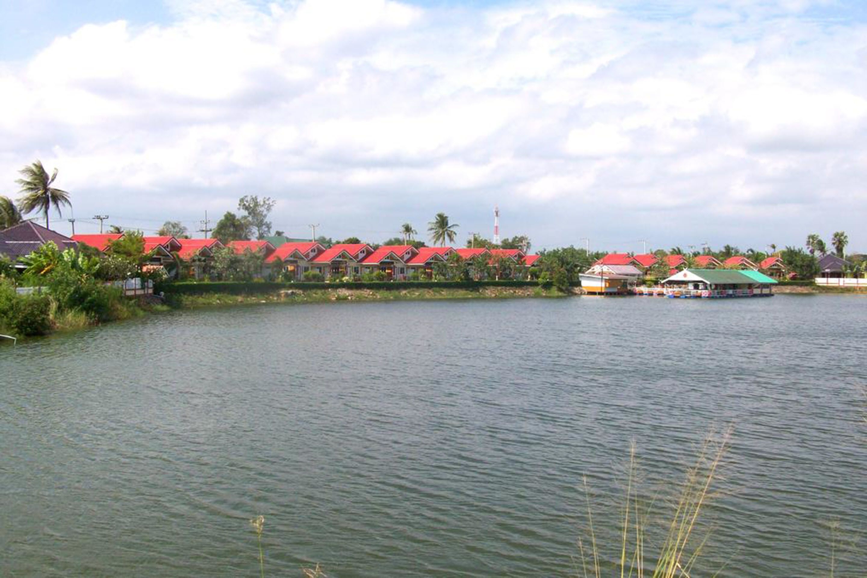 Vista de un lago con hileras de casas de techo rojo, un pequeño muelle con barcos y cielo despejado.