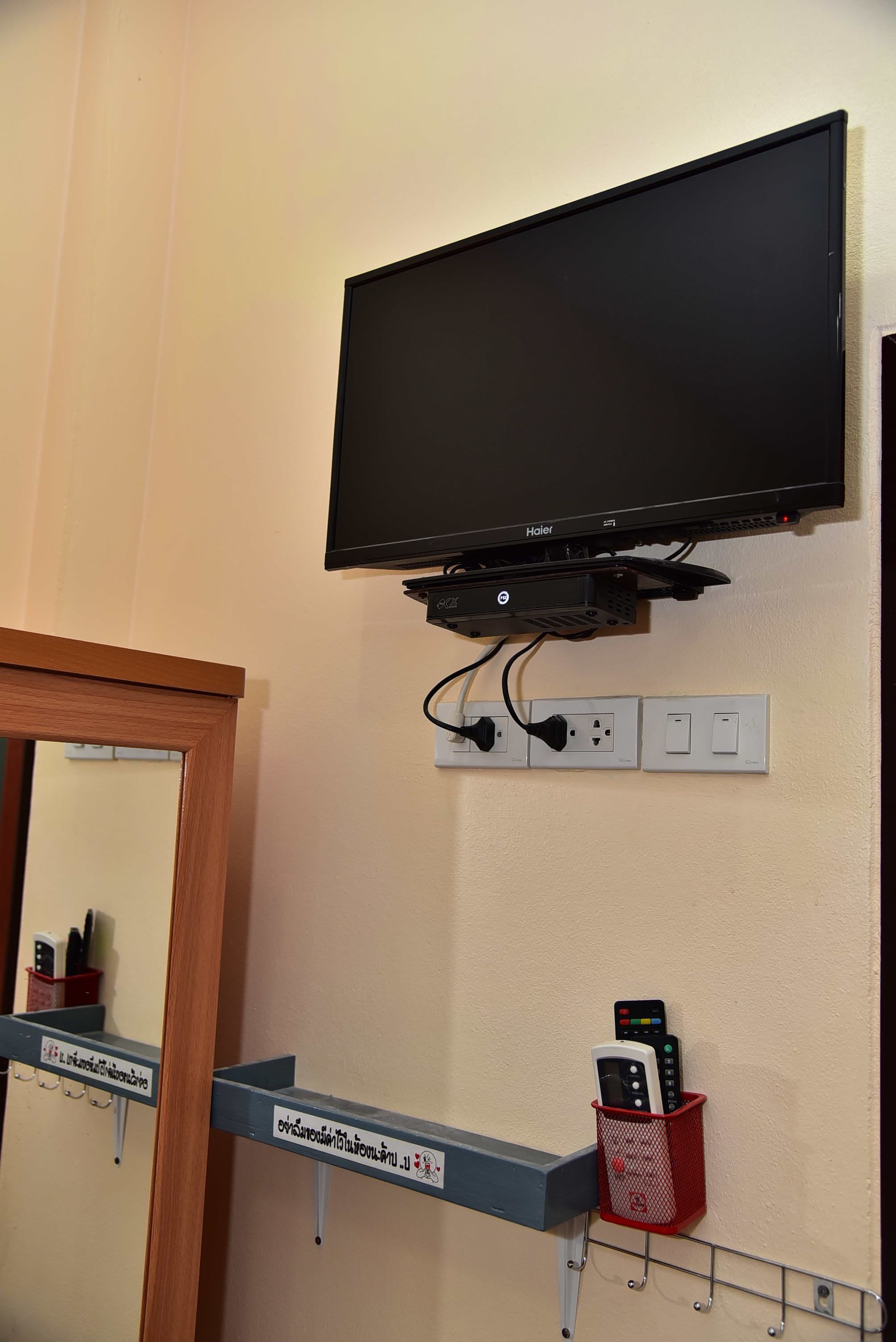 Uma televisão presa à parede, um espelho e uma prateleira com uma cesta vermelha cheia de controles remotos.
