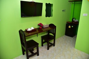 Een kamer met een groene wand, een houten tafel met twee stoelen, een televisie boven de tafel bevestigd en een kleine koelkast.