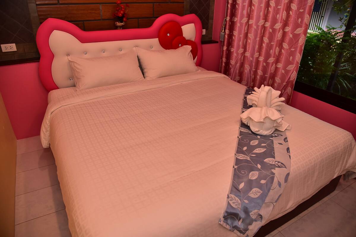 Una cama con cabecera rosa y ropa de cama blanca, una manta con estampado floral y vistas al exterior.