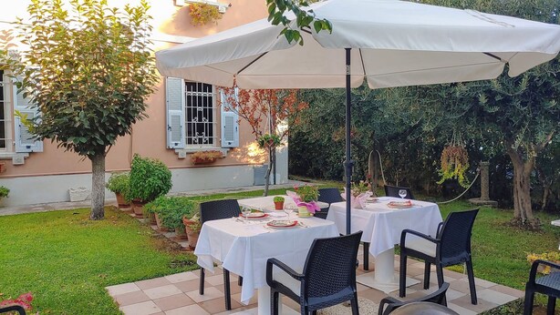 An outdoor dining area with two tables under umbrellas, surrounded by greenery and a building with shutters. - Villa Liberty B&B (Fano)