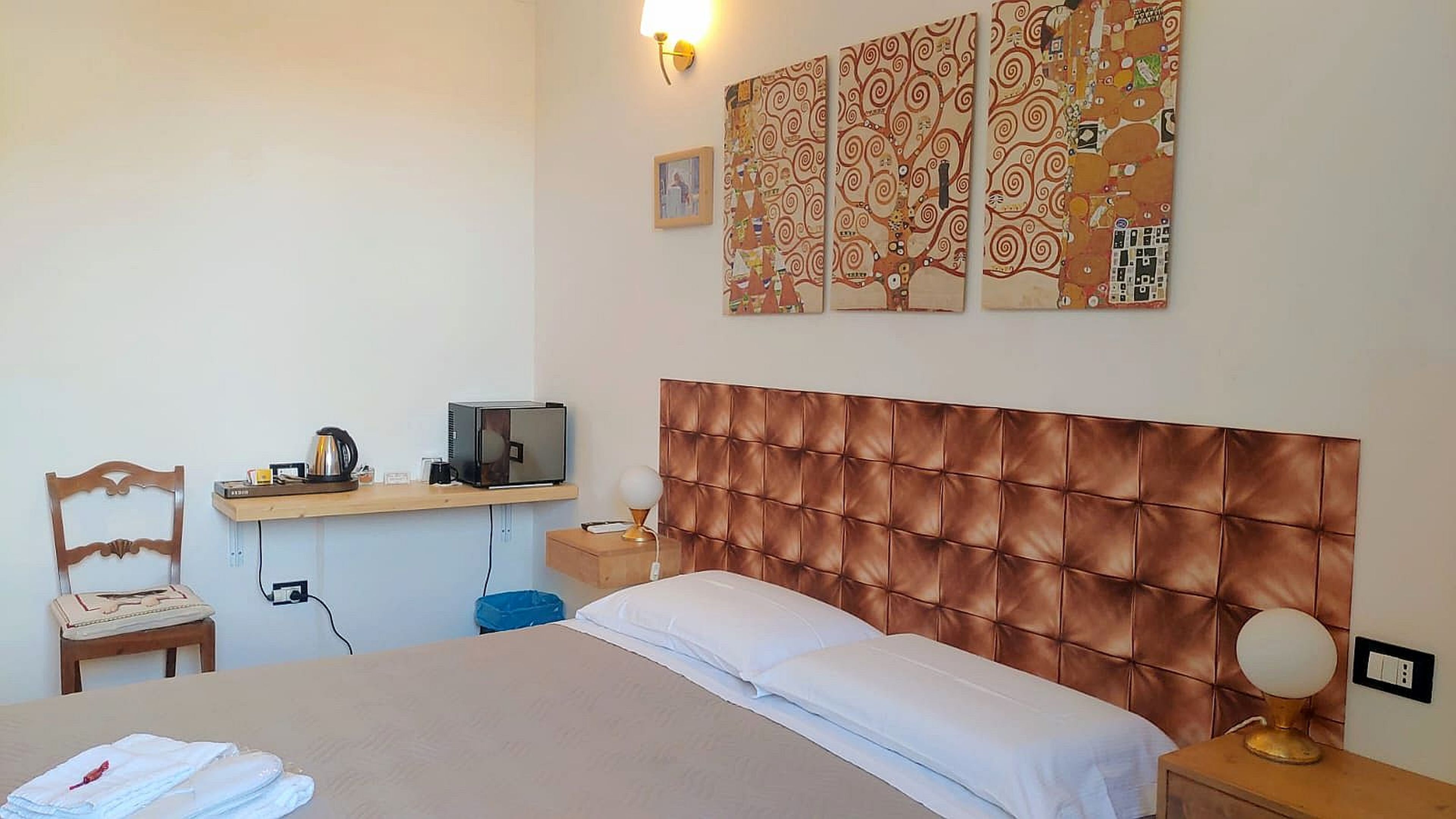 A hotel room with a bed, a desk with a computer, a chair, and a wall with artwork.