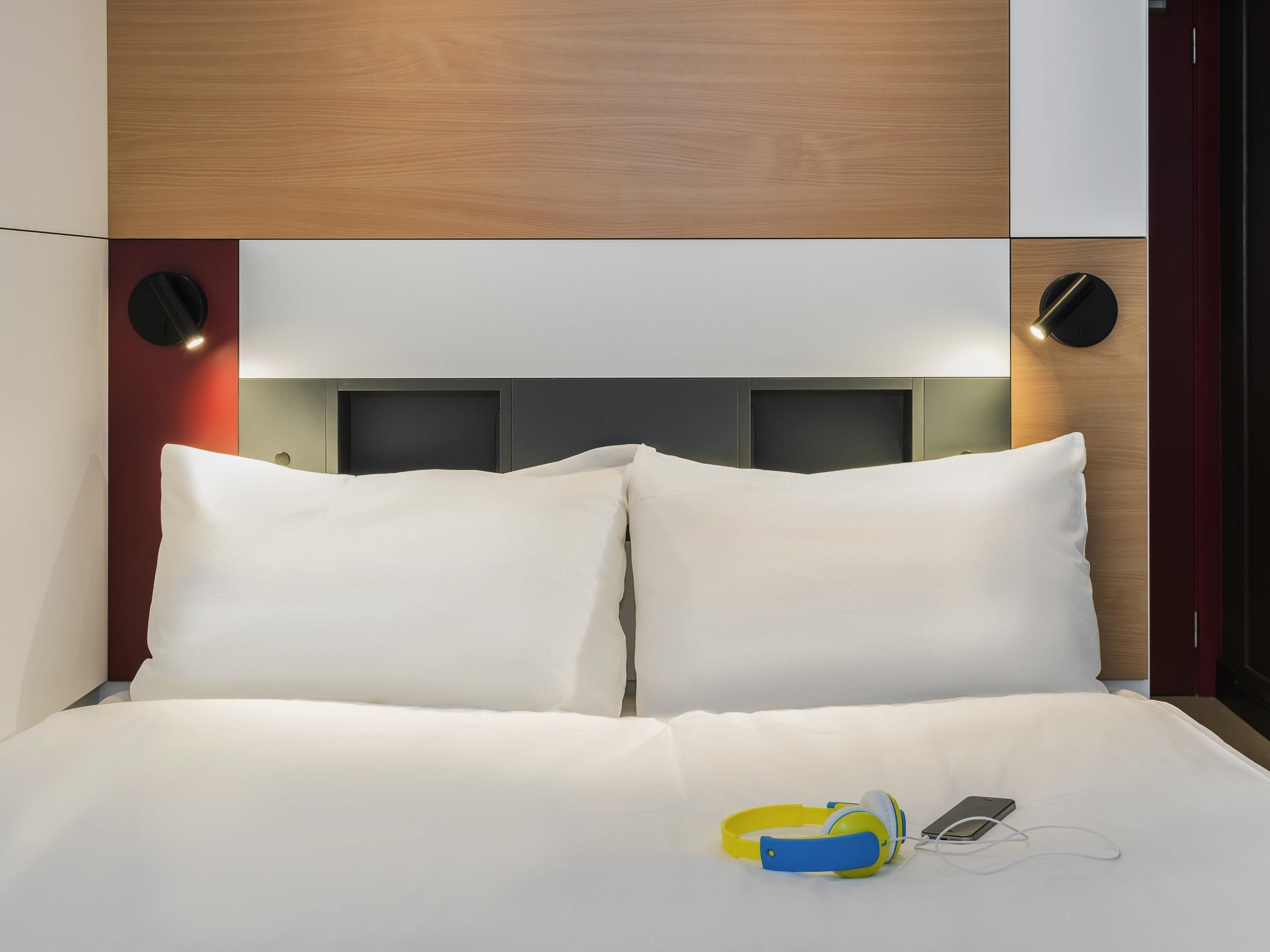 Een hotelkamer met een houten hoofdbord, twee witte kussens, een bed met een witte dekbedovertrek en een koptelefoon op het bed.