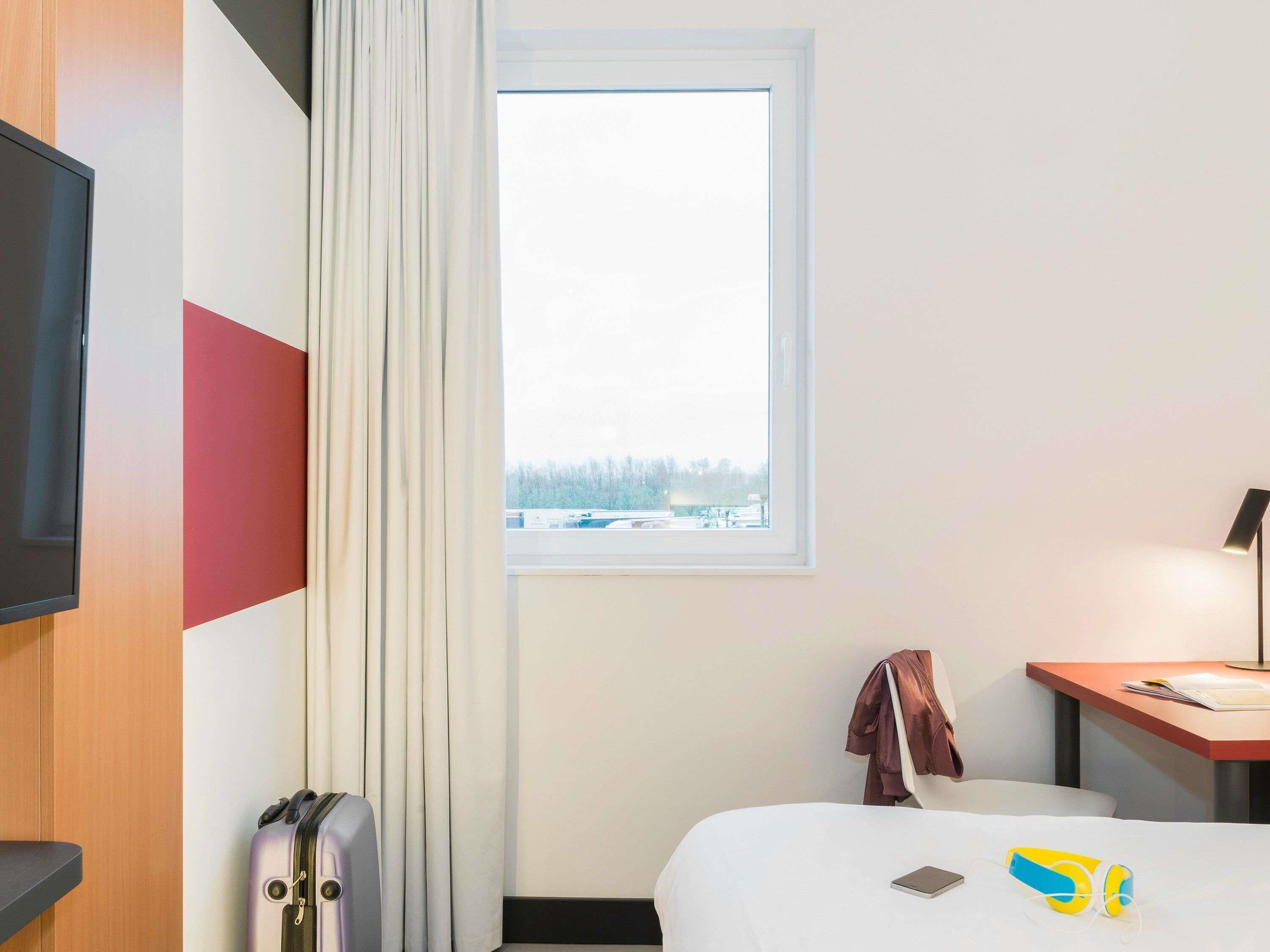 A hotel room with a bed, a desk, a television, a window, and a suitcase.