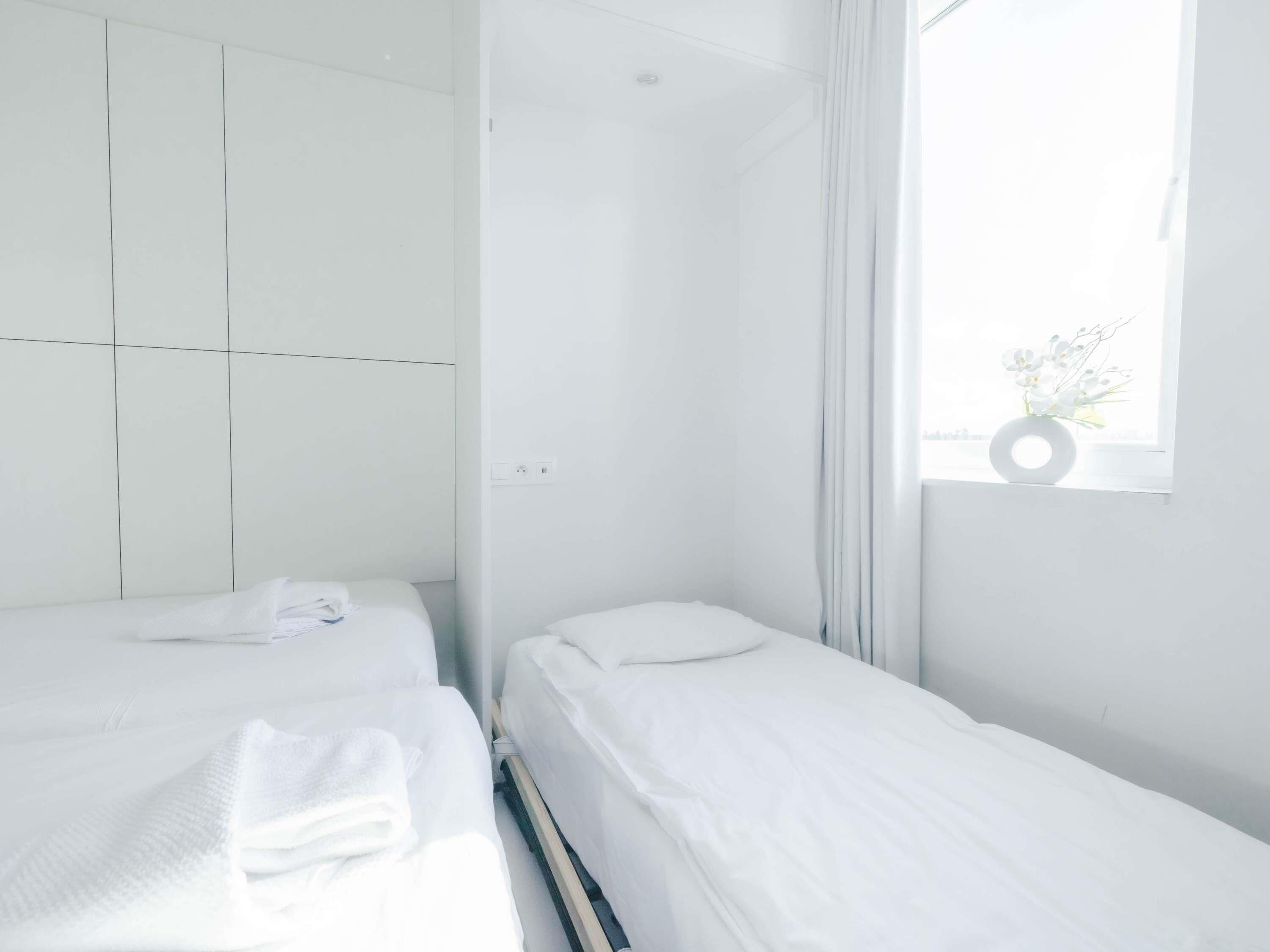 Un peque ño hotel blanco de una sola habitación con dos camas, una ventana con vistas y un florero con flores en el alféizar.