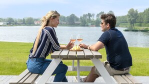 Deux personnes sont assises à une table de pique-nique en bois, avec une vue sur un lac et des maisons en arrière-plan.
