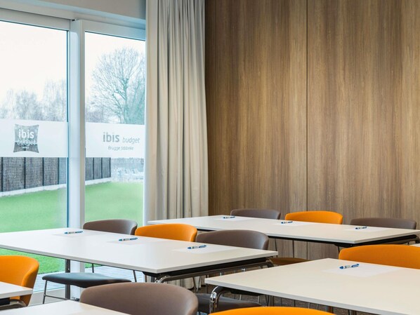 Una sala de conferencias con mesas y sillas, un ventanal con vistas a un muro de ladrillo y árboles, y un letrero que dice 'ibis budget Brugge La Pabeke'.