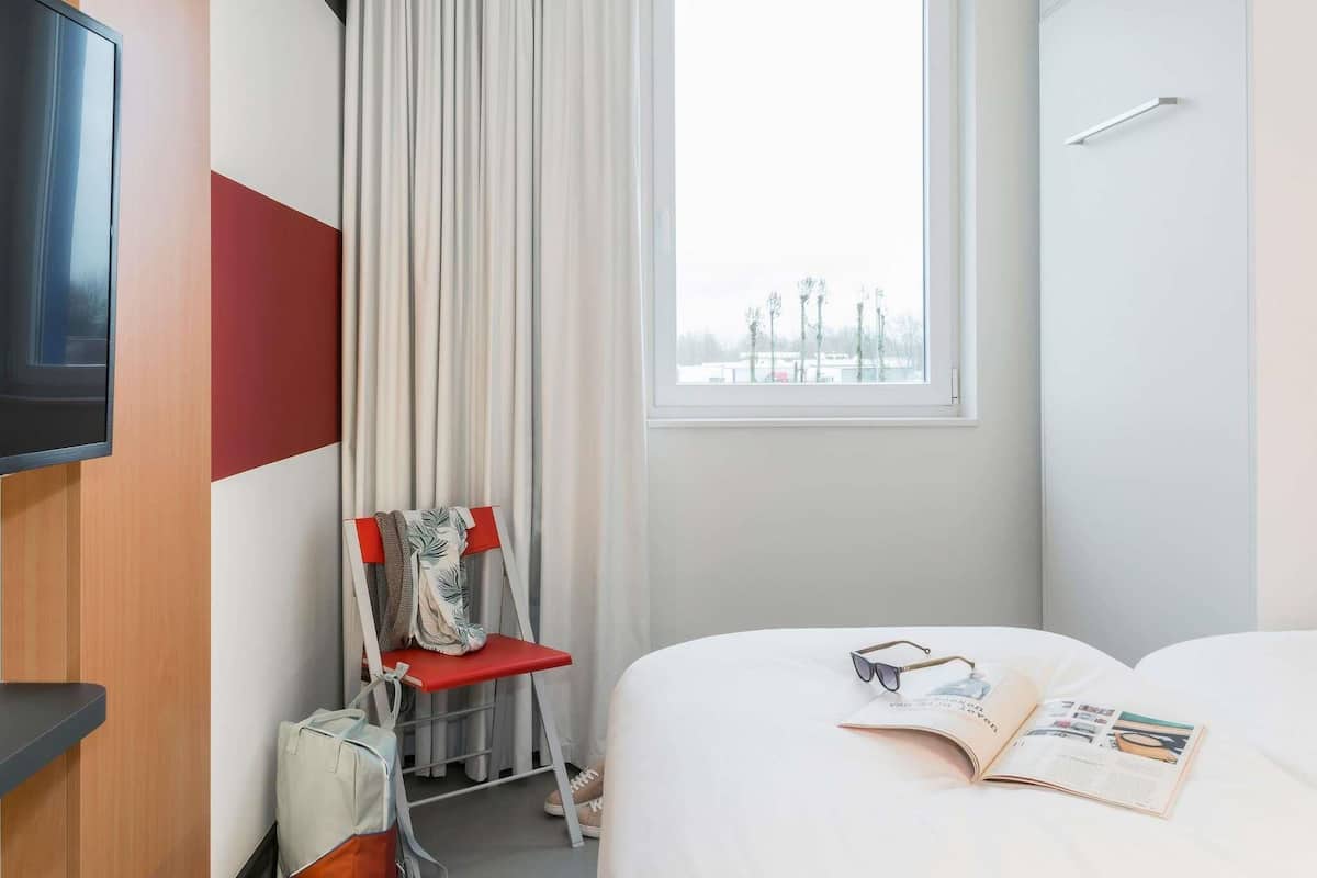 Une chambre dâhĂŽtel avec un lit, une fenĂȘtre, une chaise, un tĂ©lĂ©viseur et un magazine.