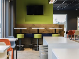 Un comedor moderno con mesas altas y taburetes, un televisor de pantalla plana en la pared y ventanas amplias que aportan luz natural.