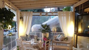 A cozy outdoor dining area with white furniture, potted plants, and a view of a slide in the background. - Hotel Loretta & Dependance (Cervia)
