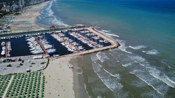 An aerial view of a coastal marina with boats docked, a beach with umbrellas, and a cityscape in the background. - Hotel Loretta & Dependance (Cervia)