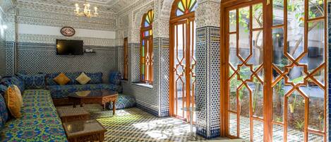 Quarto com trabalhos em azulejos detalhados, sofá, televisão e grandes janelas com vidros decorativos.