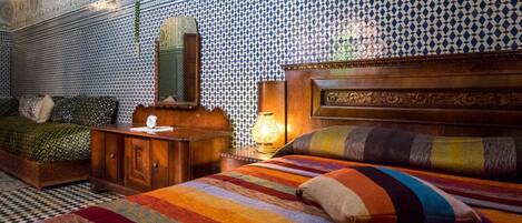 Una habitación con una cama, una mesita de noche de madera, una lámpara y un espejo. Las paredes tienen un diseño de azulejos estampados.