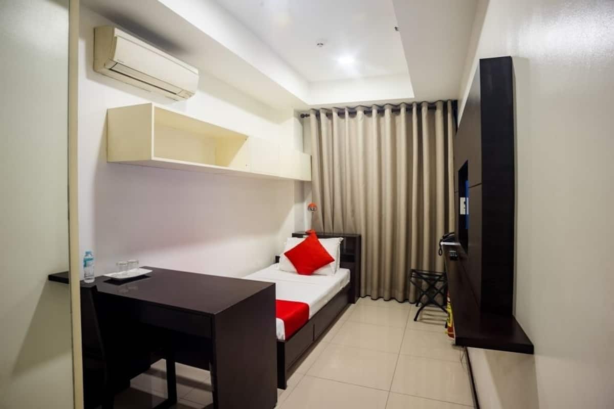 A compact hotel room with a bed, a desk, a TV, and an air conditioning unit.