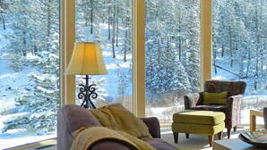 A cozy room with a large window overlooking a snowy forest. There is a sofa, an ottoman, and a chair with a patterned cushion. A table with a tray is visible in the foreground.