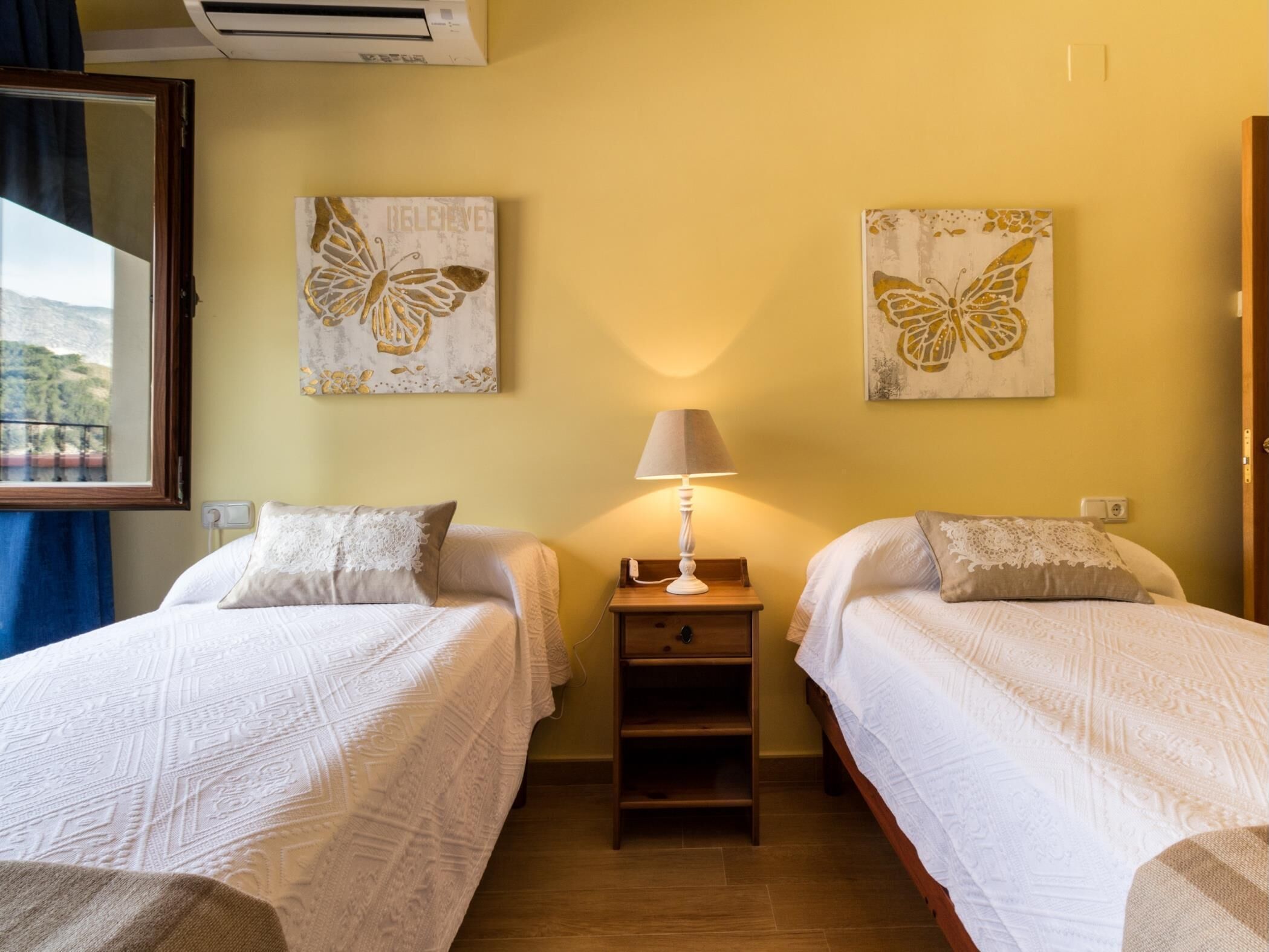 A hotel room with two beds, a nightstand, a lamp, and two butterfly-themed wall art pieces.