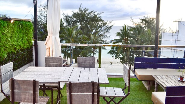 An outdoor seating area with wooden tables and chairs, a white umbrella, and a view of the beach and palm trees.