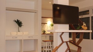 A modern room with a TV mounted on a wooden stand, a small seating area with a plant, and a kitchenette in the background.