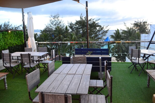 An outdoor patio with wooden tables and chairs, surrounded by green artificial turf and palm trees, overlooking a body of water.