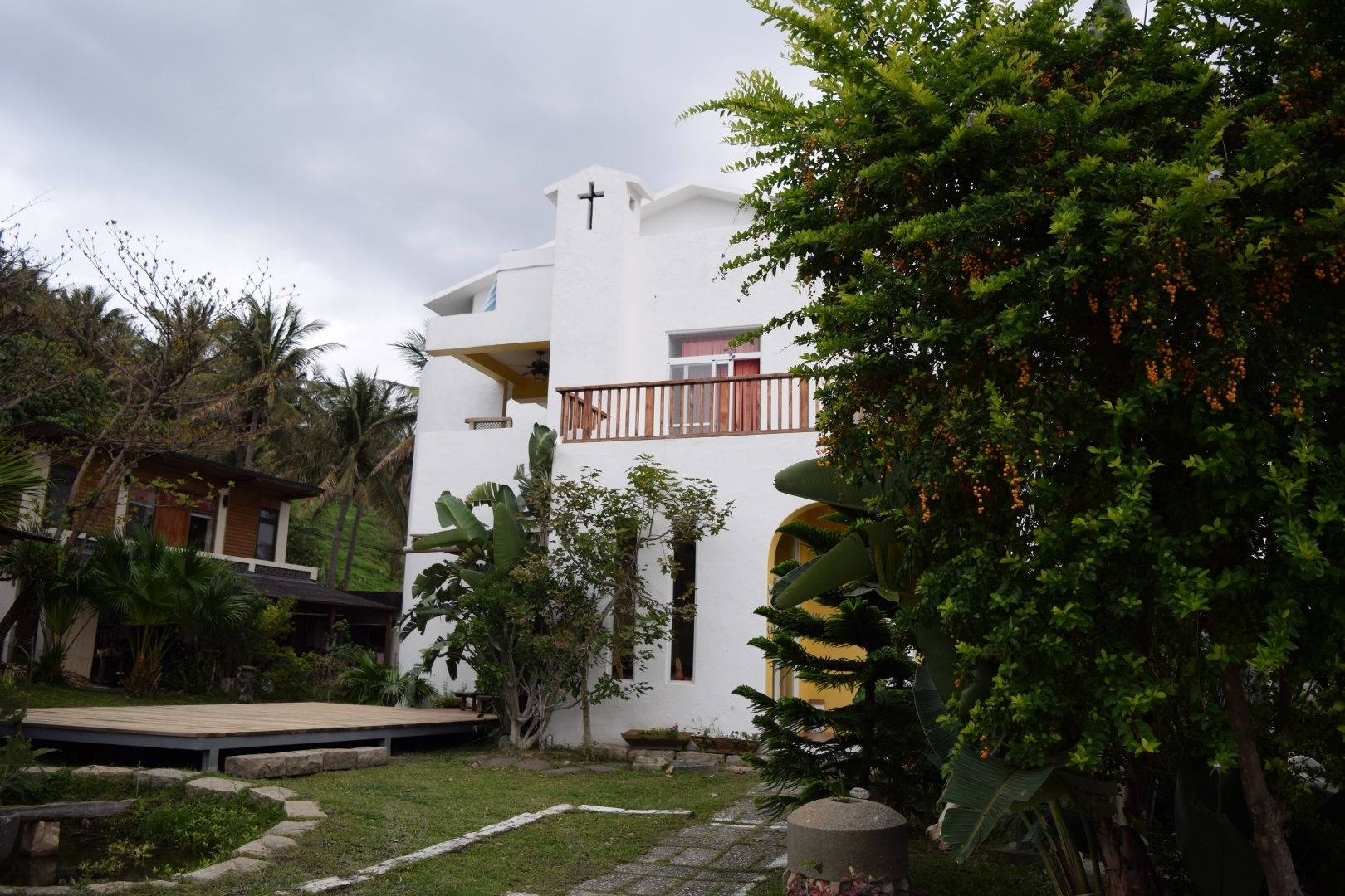 A white building with a cross on top, surrounded by lush greenery and a wooden deck.