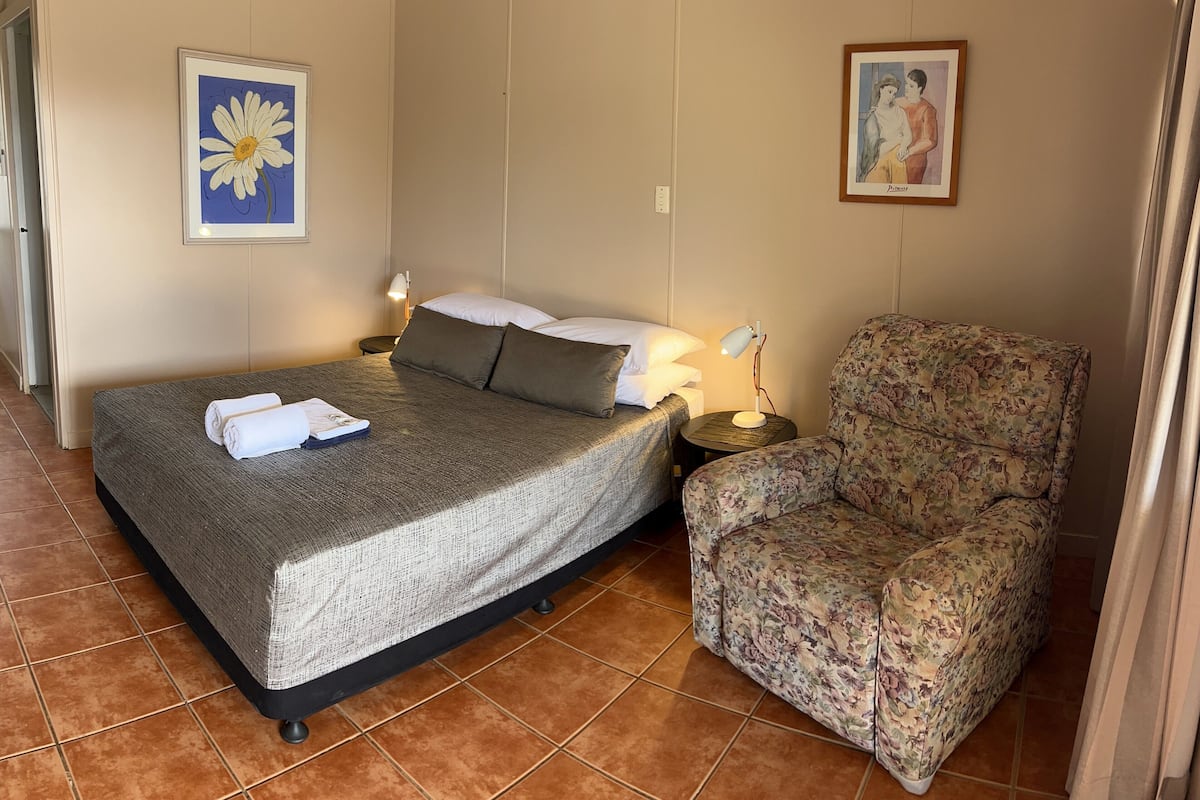 Habitación de hotel con cama, una silla, mesita de noche, lámpara y dos cuadros enmarcados en las paredes.