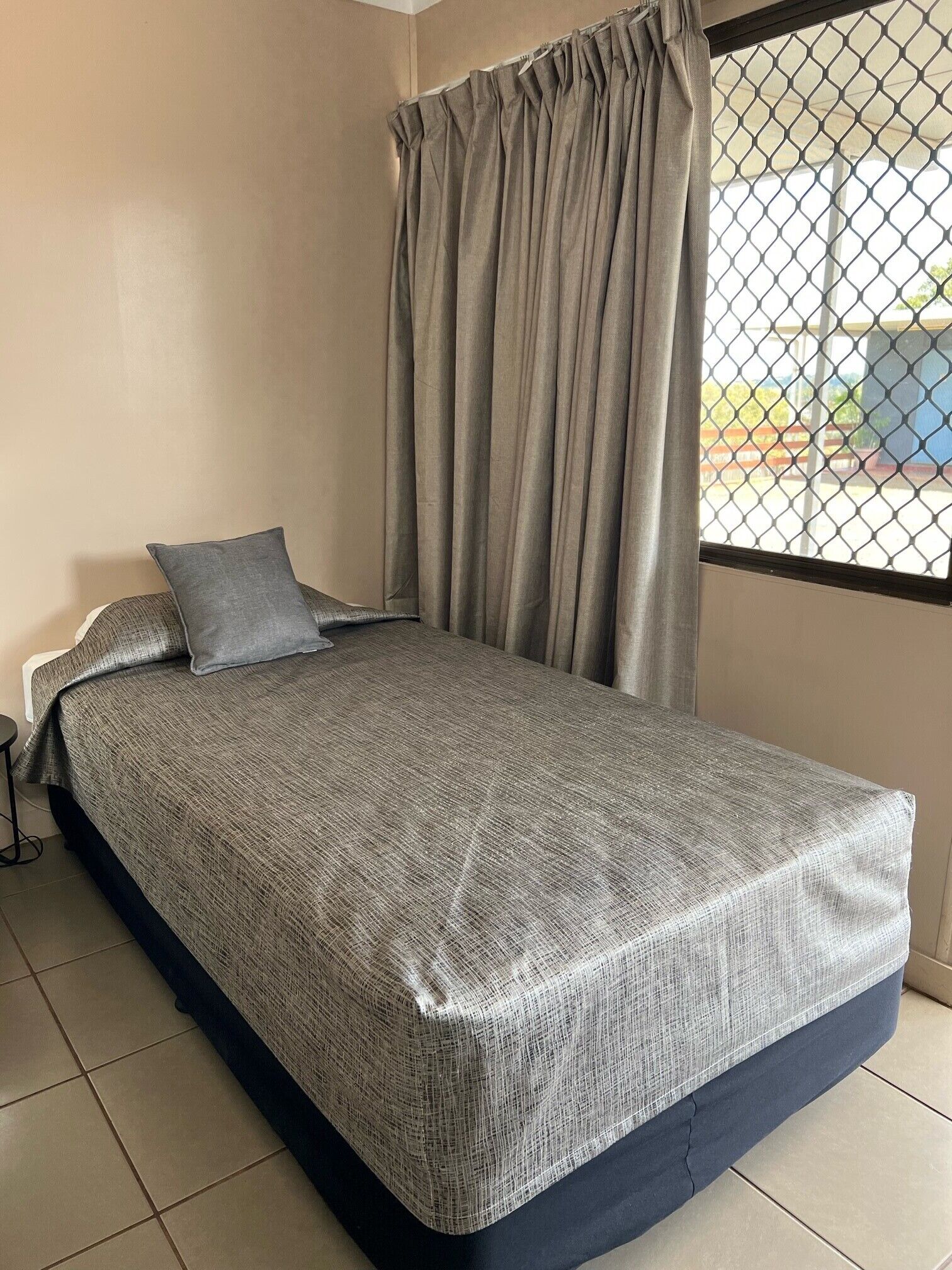 A single bed with a grey bedspread, a grey pillow, and a window with a metal screen.