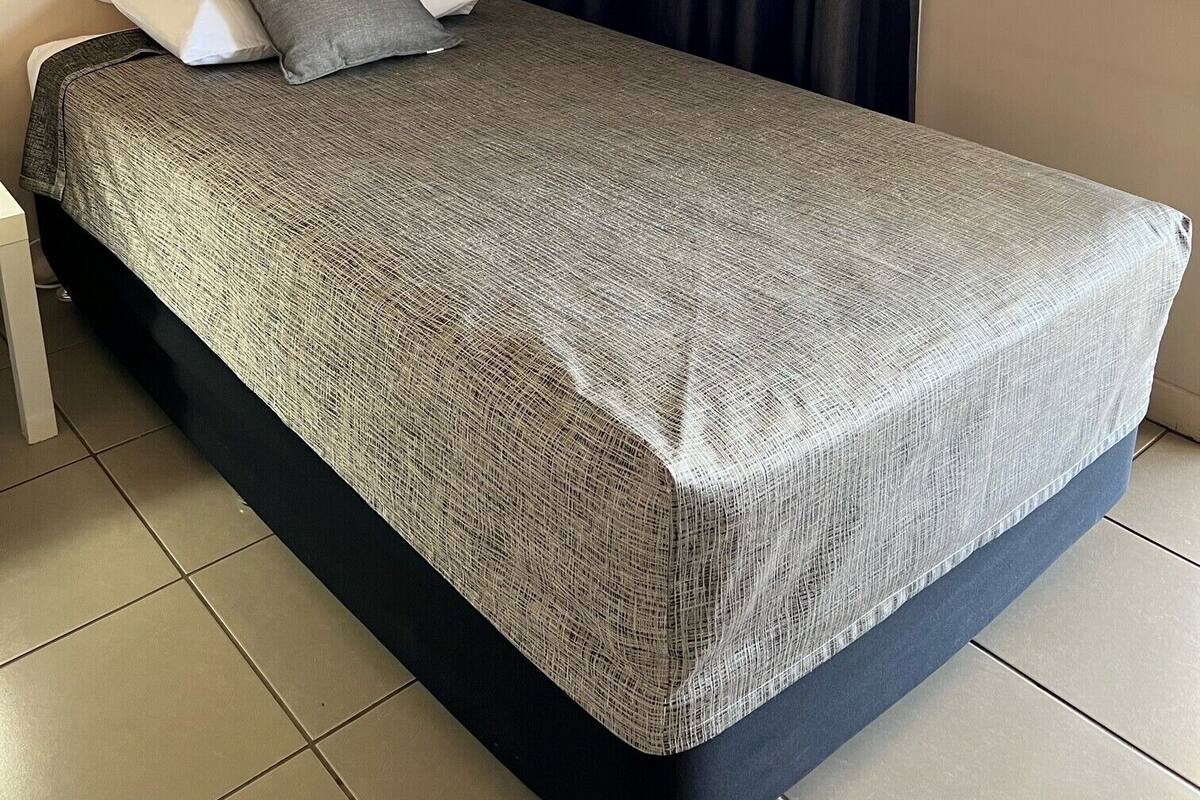 Una cama individual con una colcha estampada, dos almohadas, una cortina oscura y una ventana con mosquitero.