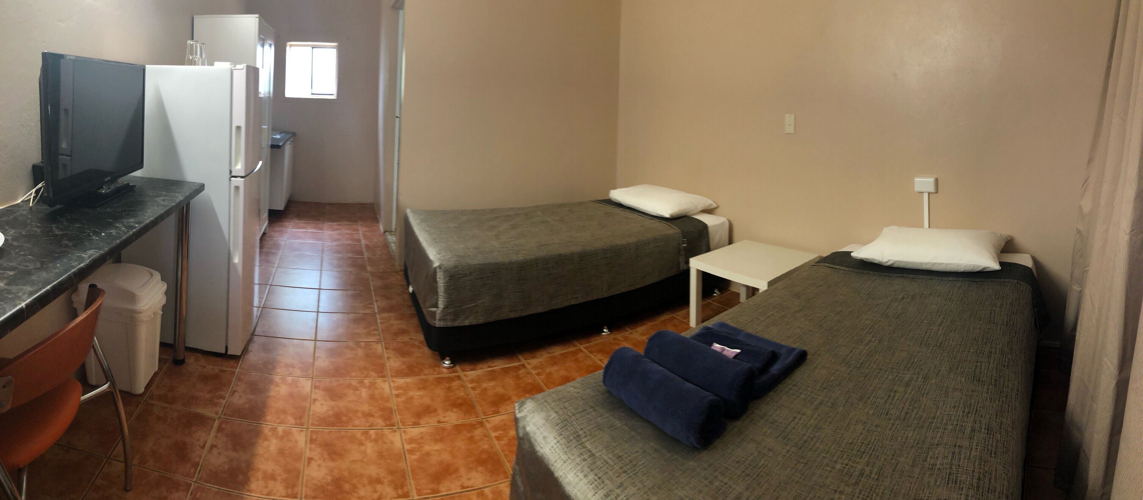Une chambre d’hôtel compacte comprenant deux lits simples, un petit bureau avec un ordinateur et un coin cuisine.