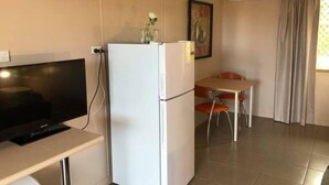 A compact room with a flat-screen TV, a white refrigerator, a small table with two chairs, and an air conditioning unit. - Childers Budget Accommodation (Childers)