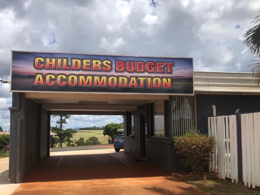 Une enseigne indiquant « CHILDERS BUDGET ACCOMMODATION », avec en arrière-plan un parking et l’entrée d’un immeuble.