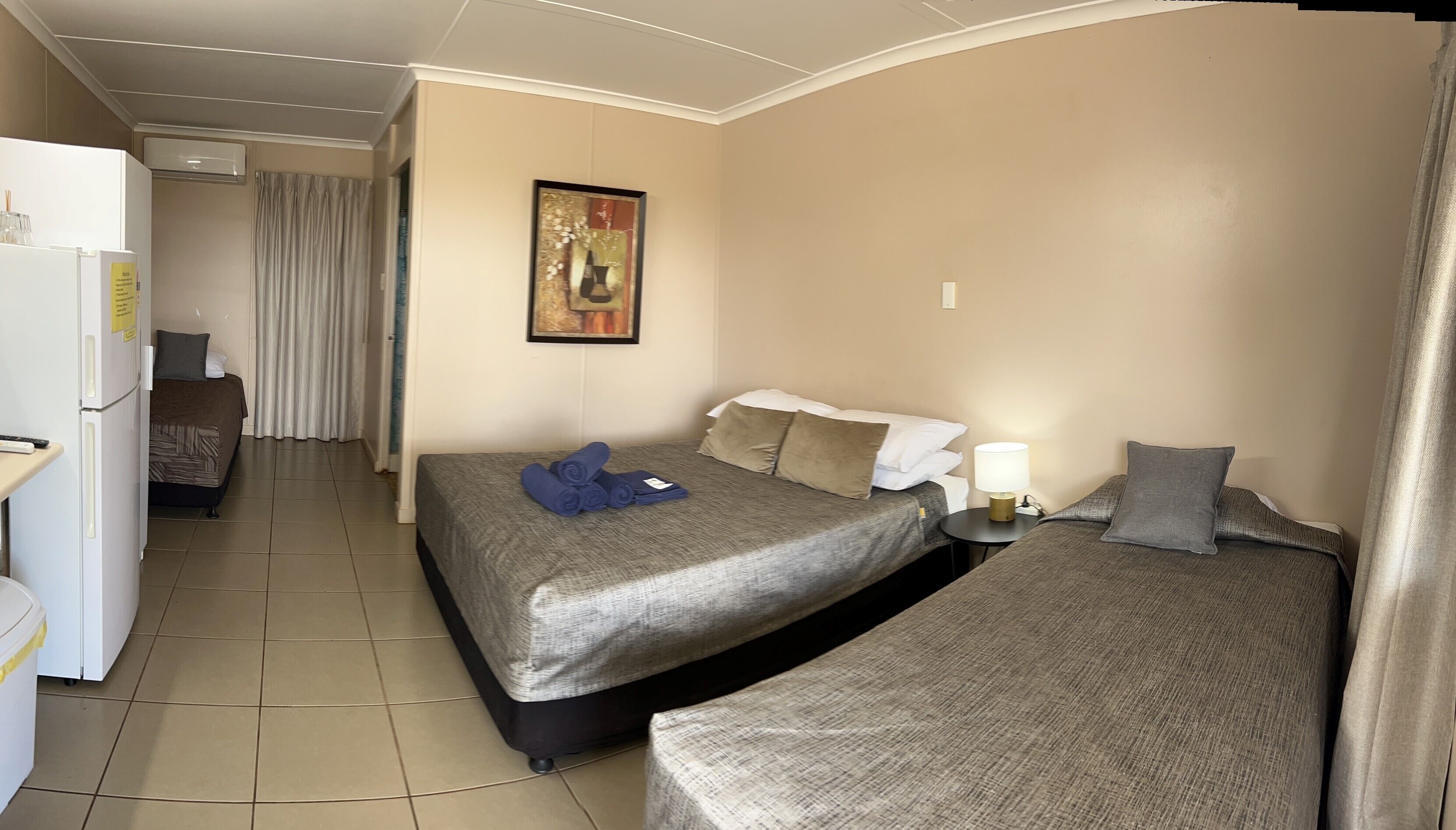 A hotel room with two beds, a refrigerator, a small table with a lamp, and a painting on the wall.