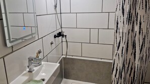 Un baño con paredes de azulejos blancos, un lavabo con una toalla verde, un cabezal de ducha de mano y un espejo.
